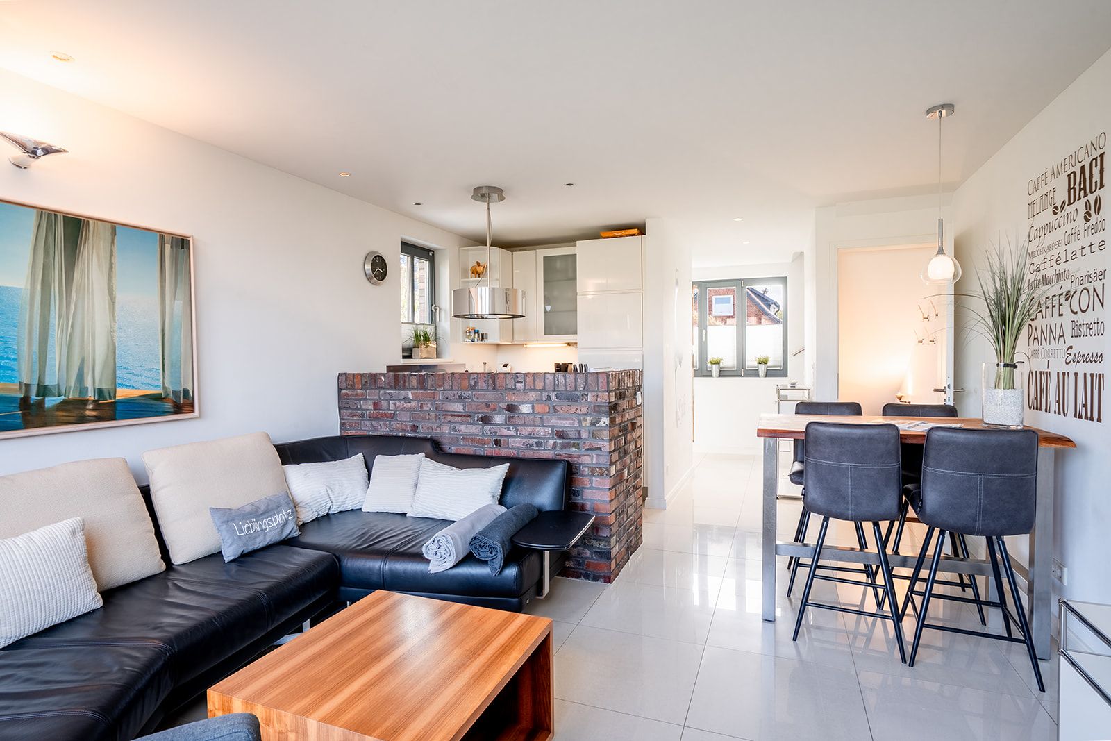 Helles Wohnzimmer mit schwarzem Sofa, Holztisch, offener Küche und Essbereich mit vier Stühlen.