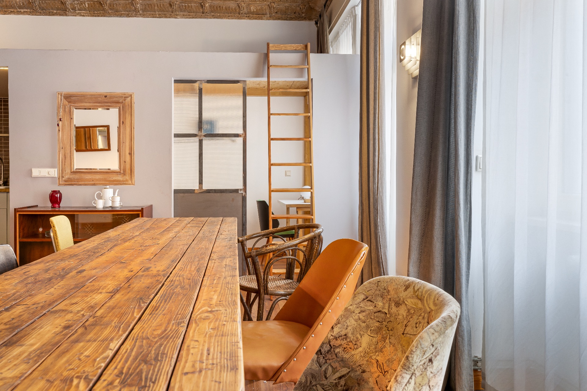 Wohnzimmer mit Holztisch, verschiedenen Stühlen, Leiter und großen Fenstern mit Vorhängen.