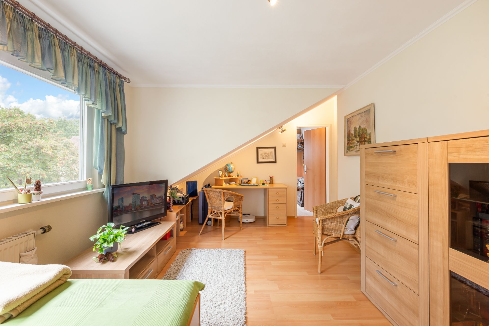 Helles Wohnzimmer mit Schreibtisch, TV, Holzschrank und großem Fenster mit Blick ins Grüne.