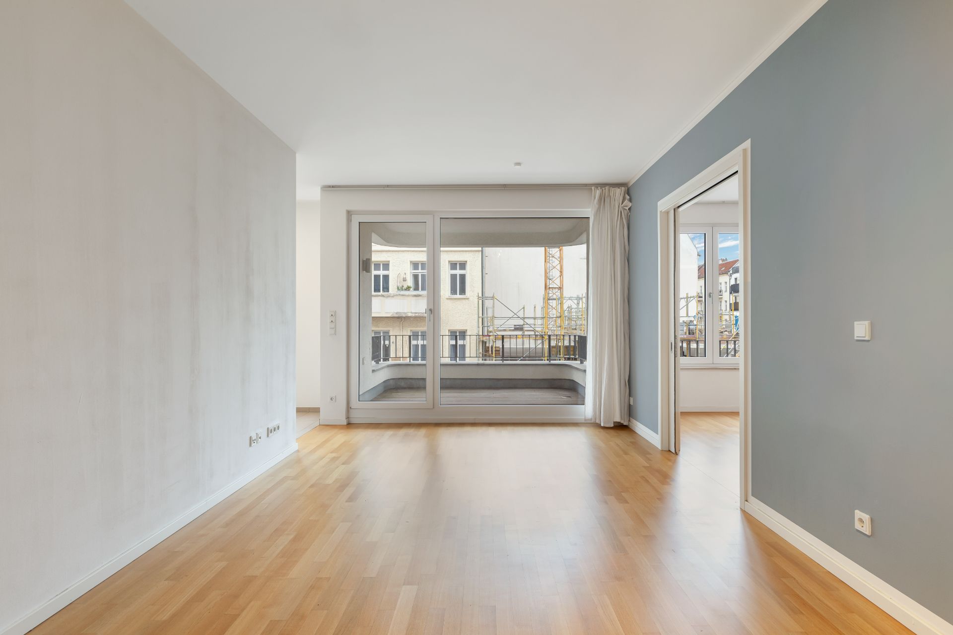 Leeres Wohnzimmer mit Holzboden, großer Fensterfront und Zugang zu einem weiteren Raum.