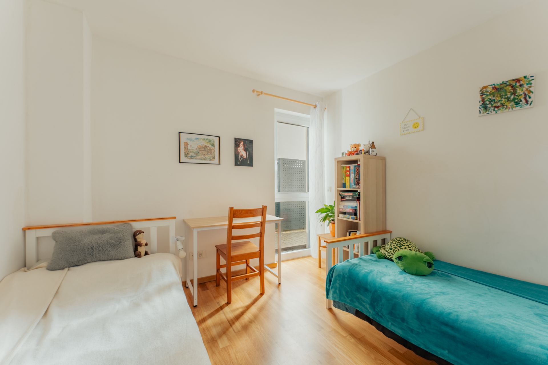 Helles Schlafzimmer mit zwei Betten, Schreibtisch, Stuhl und Regal, Holzboden und Dekoration an den Wänden.