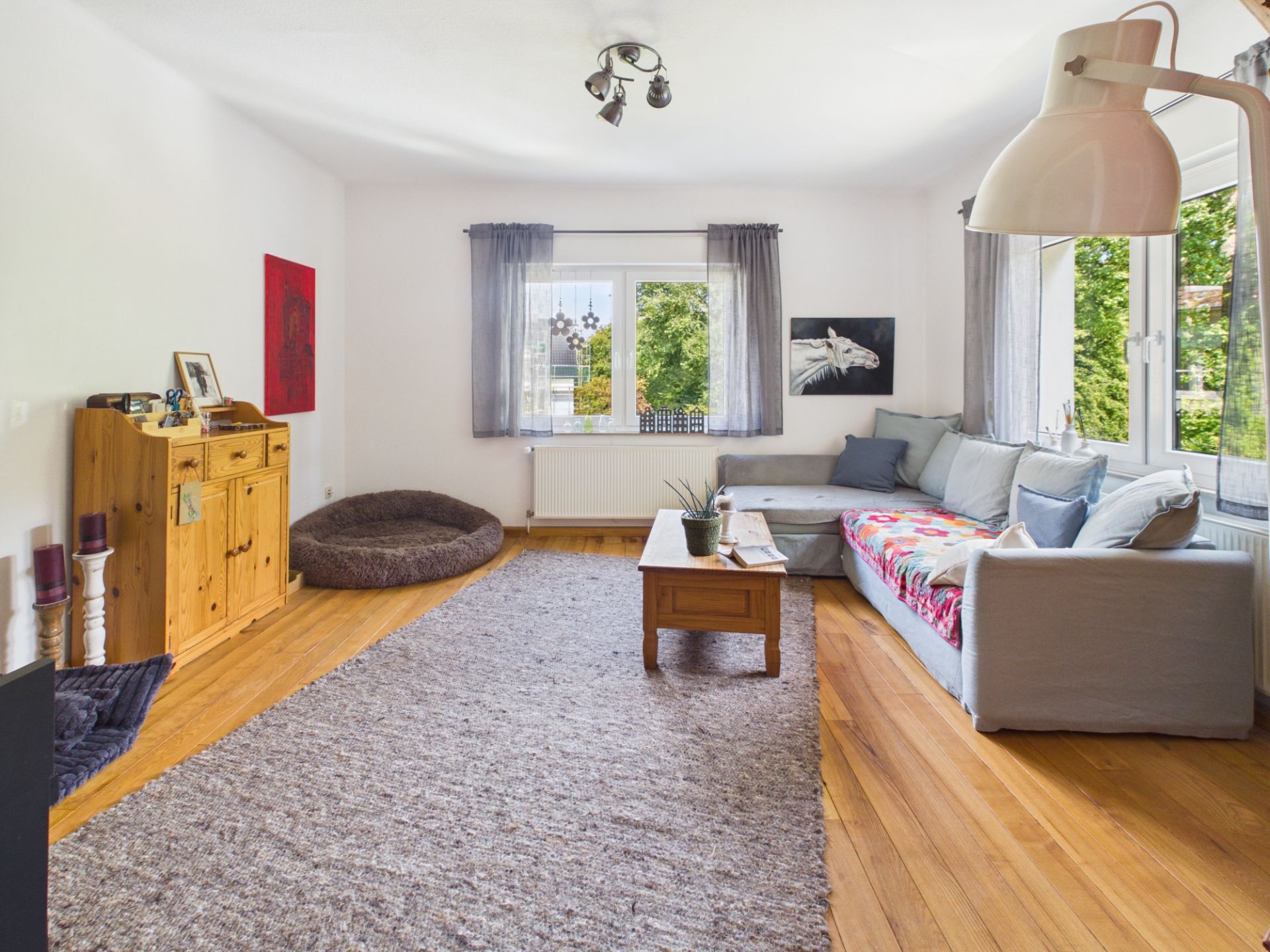 Helles Wohnzimmer mit großem Ecksofa, Holzmöbeln, Teppich und Fenstern mit Blick ins Grüne.