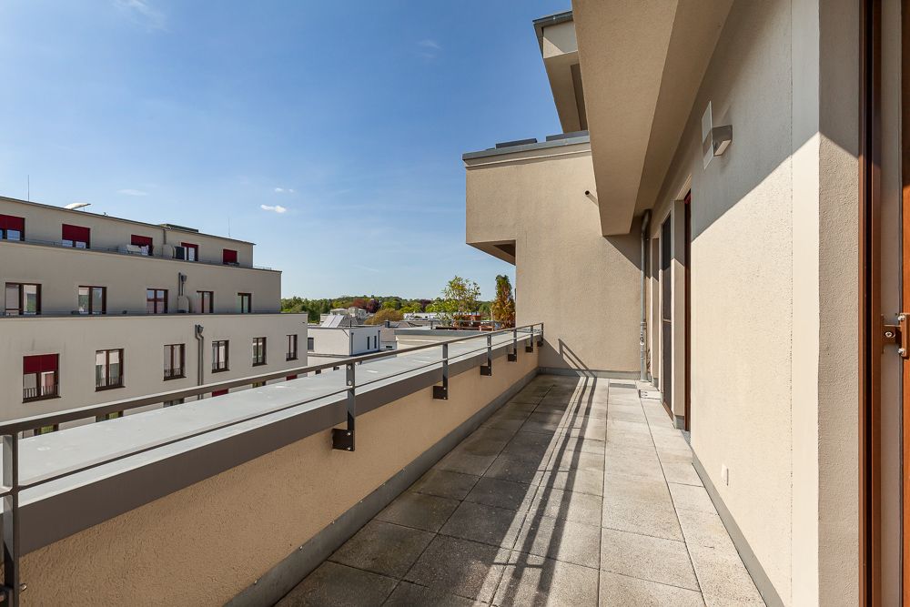 Großer Balkon mit Geländer, Blick auf benachbarte Wohngebäude und blauen Himmel.