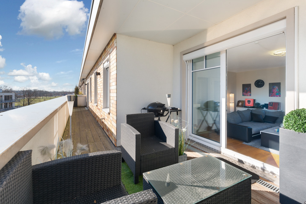 Großer Balkon mit Sitzmöbeln und Tisch, Zugang zum Wohnzimmer durch eine Glastür.