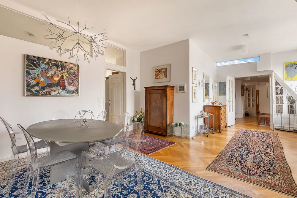 Helles Esszimmer mit rundem Tisch, transparenten Stühlen und Teppichen auf Holzboden.