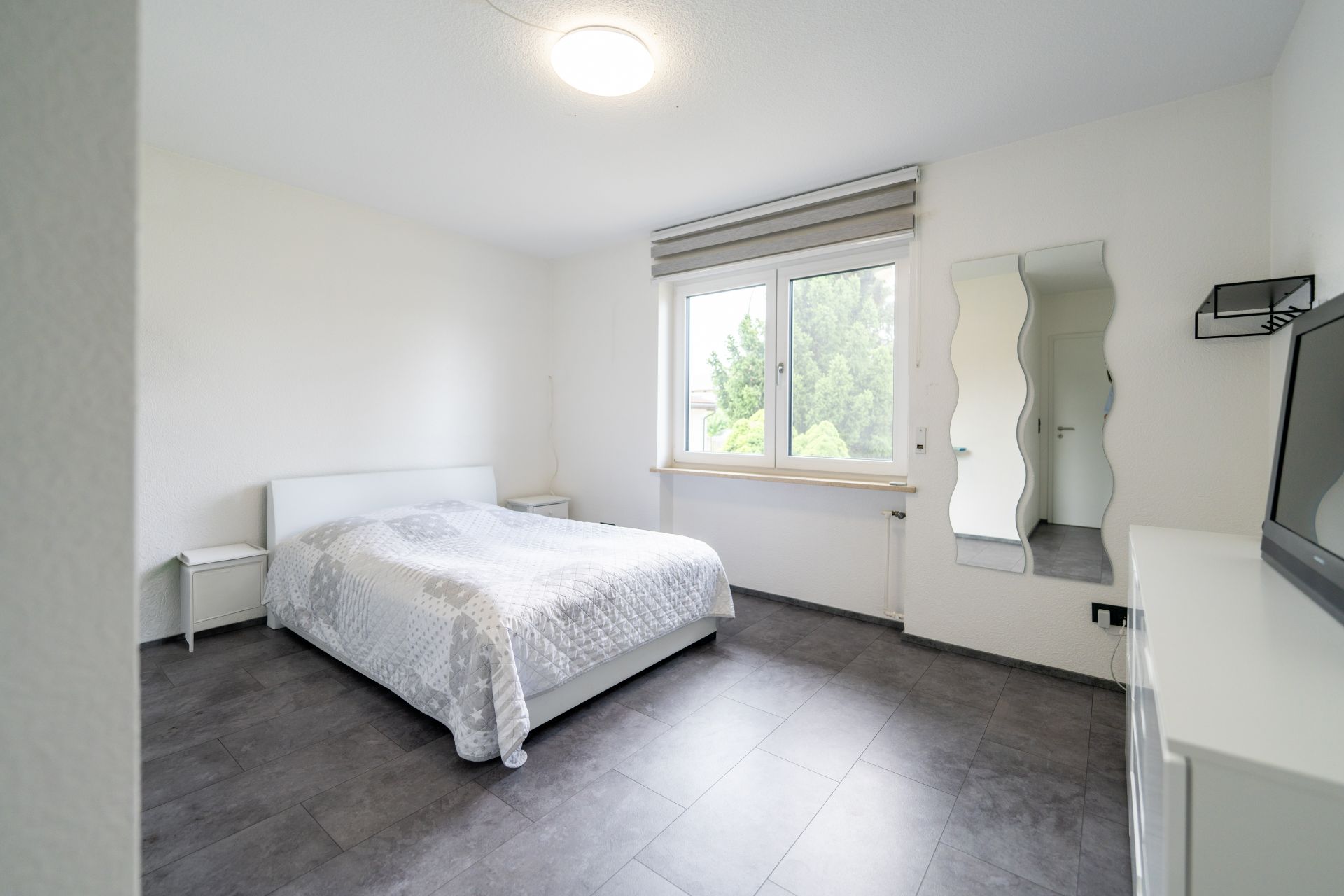 Helles Schlafzimmer mit Doppelbett, Fenster, Kommode, Spiegel und Nachttisch.