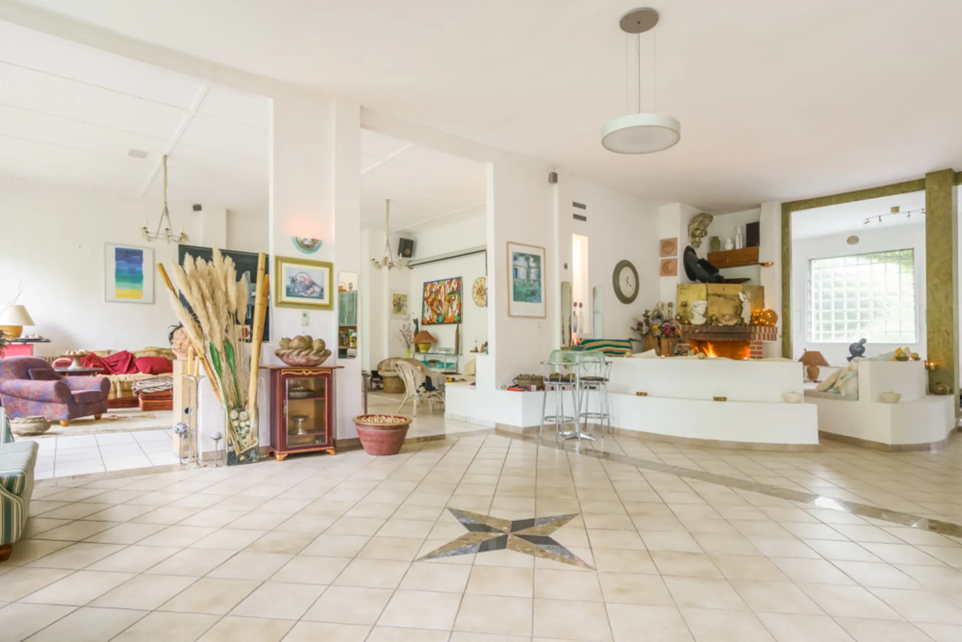 Helles Wohnzimmer mit Kunstwerken, Kamin, Barhockern und dekorativem Sternmuster auf dem Fliesenboden.