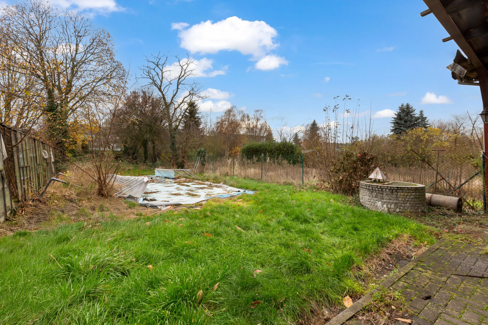 Garten mit grünem Rasen, altem Brunnen und abgedeckter Fläche, Bäume im Hintergrund.