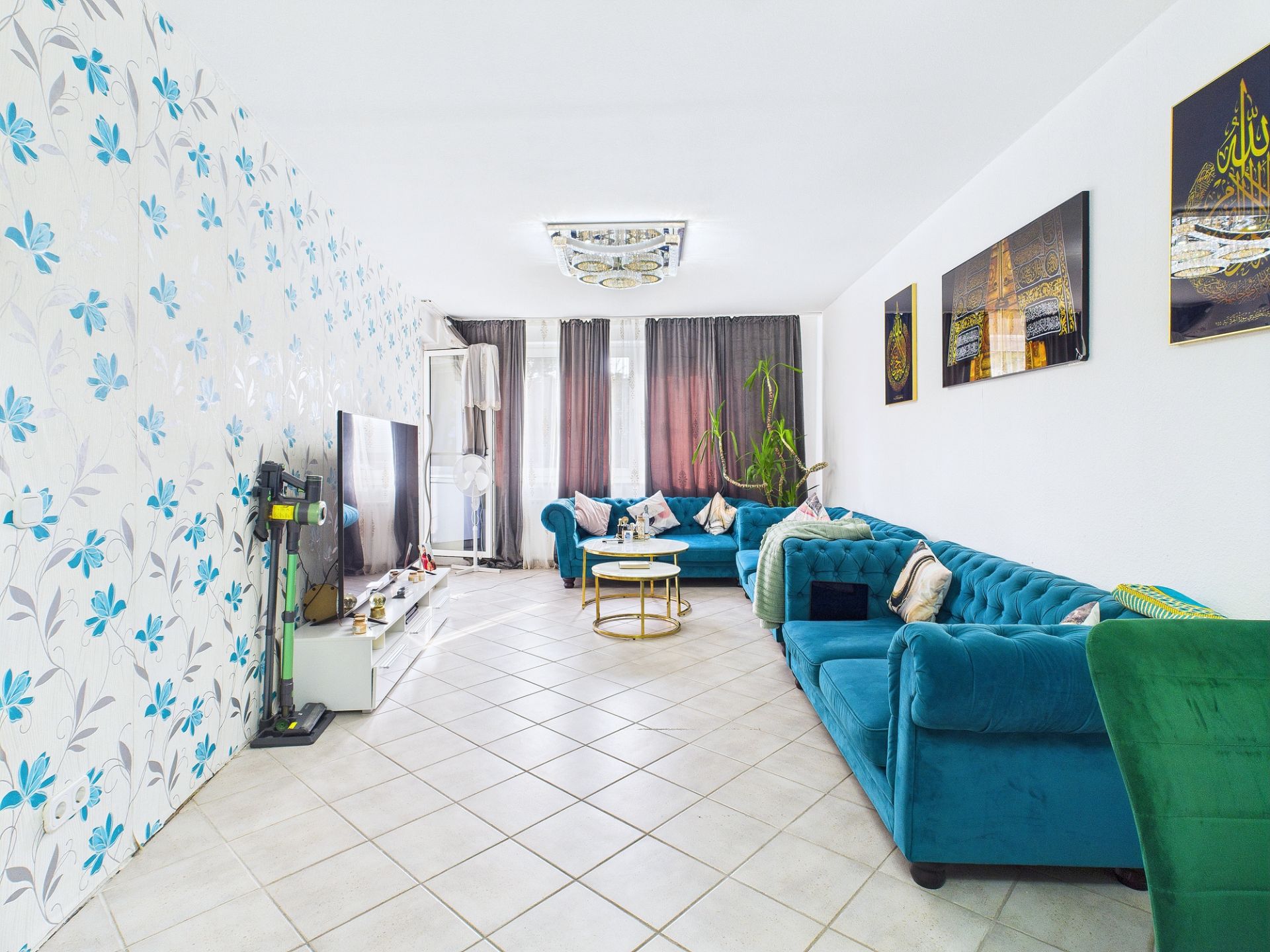 Wohnzimmer mit blauer Couch, gemusterten Kissen, Fernseher, Blumenwand und Fliesenboden.