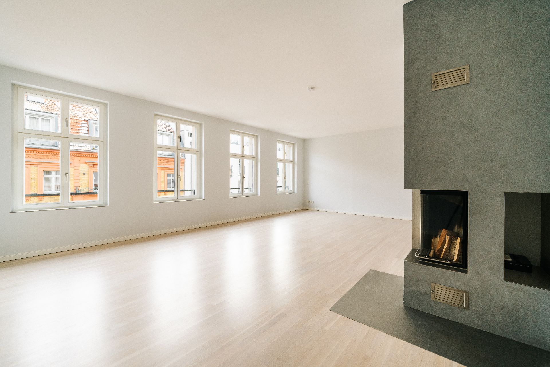 Helles, leeres Wohnzimmer mit Holzboden, großen Fenstern und modernem Kamin.
