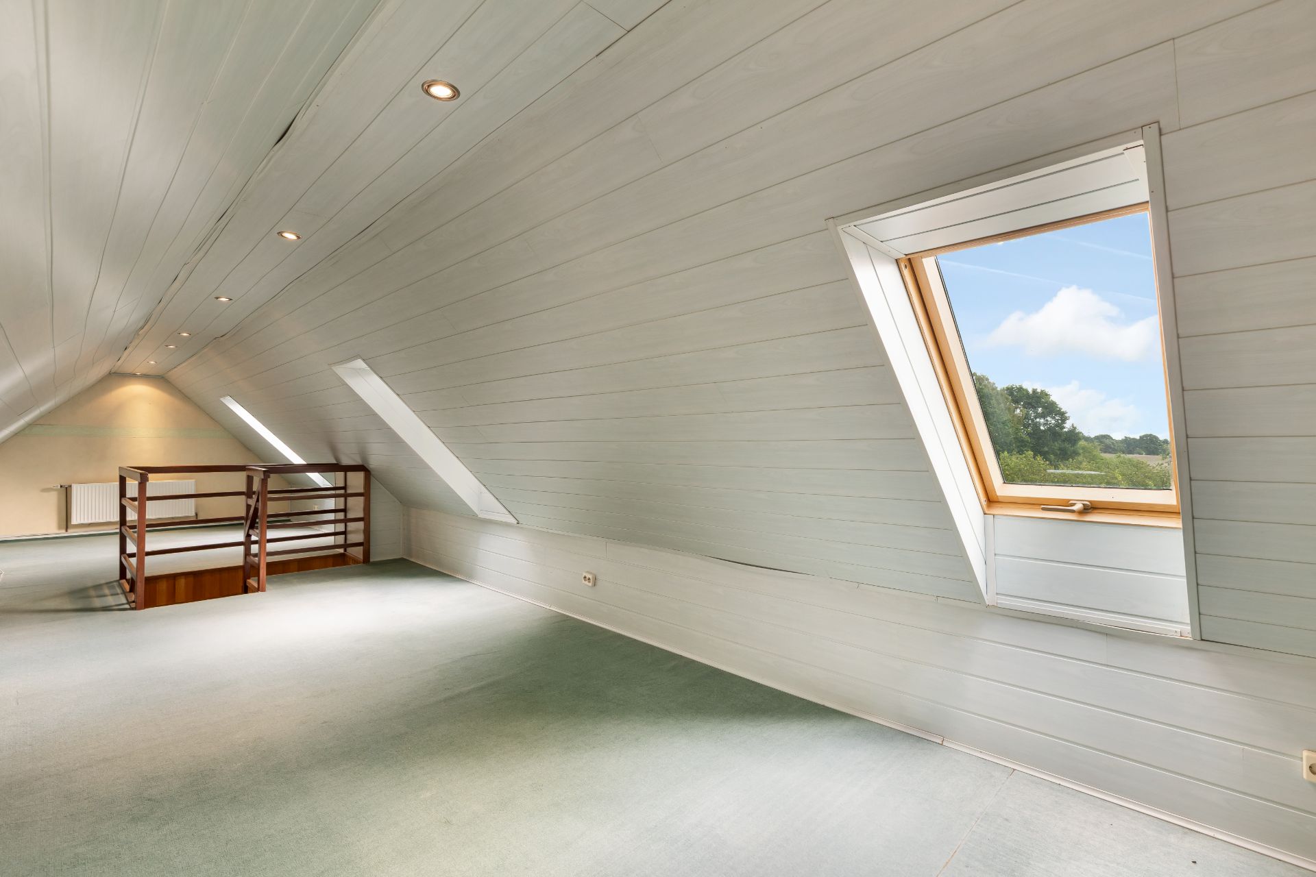 Heller Dachbodenraum mit Teppichboden, Holzgeländer und Dachfenstern mit Blick ins Freie.