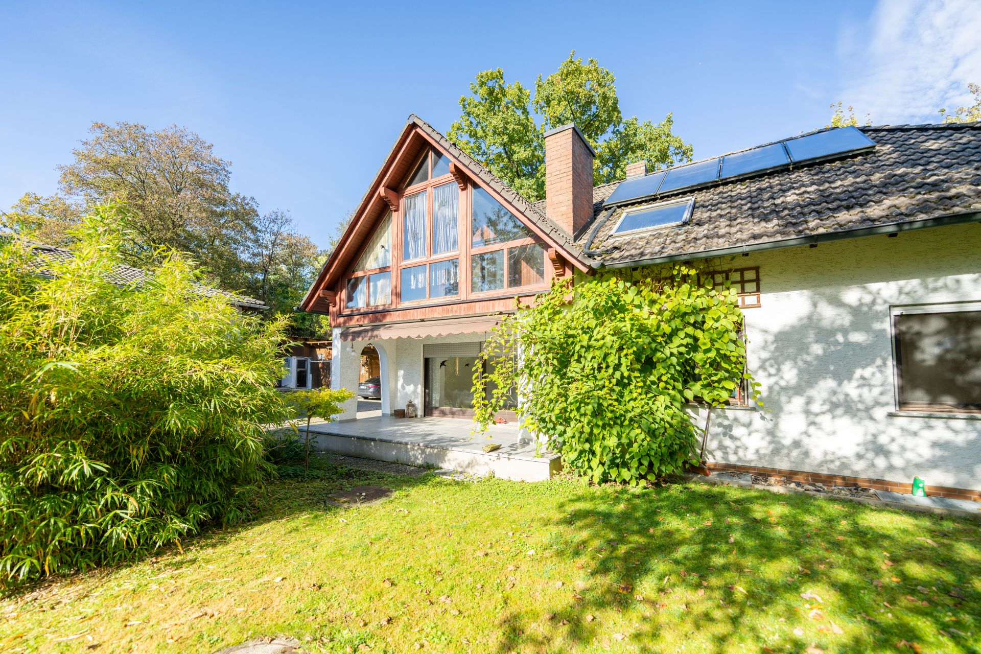 Haus mit großer Fensterfront, Balkon und Terrasse, umgeben von Garten und Bäumen.