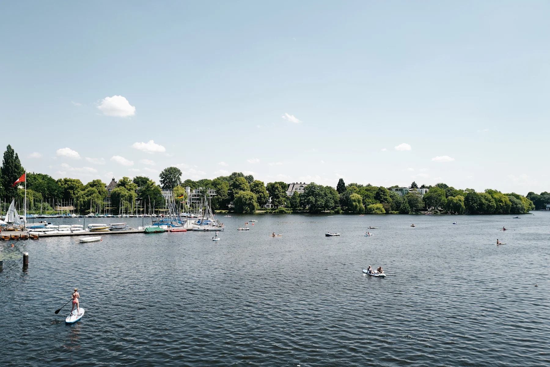 Blick auf einen See mit Segelbooten und Menschen auf dem Wasser, im Hintergrund grüne Bäume.