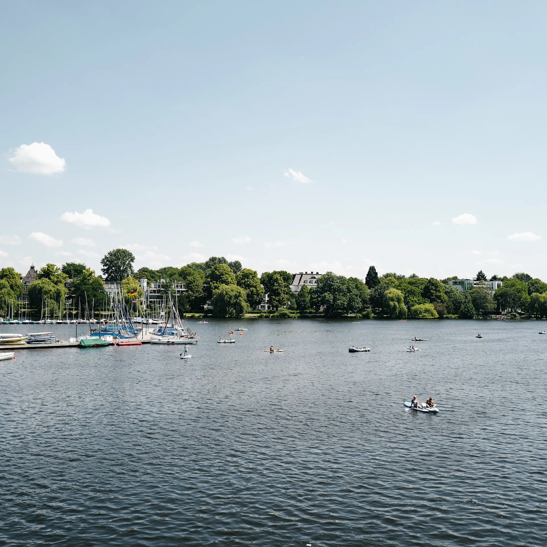 Blick auf einen See mit Segelbooten und Menschen auf dem Wasser, im Hintergrund grüne Bäume.
