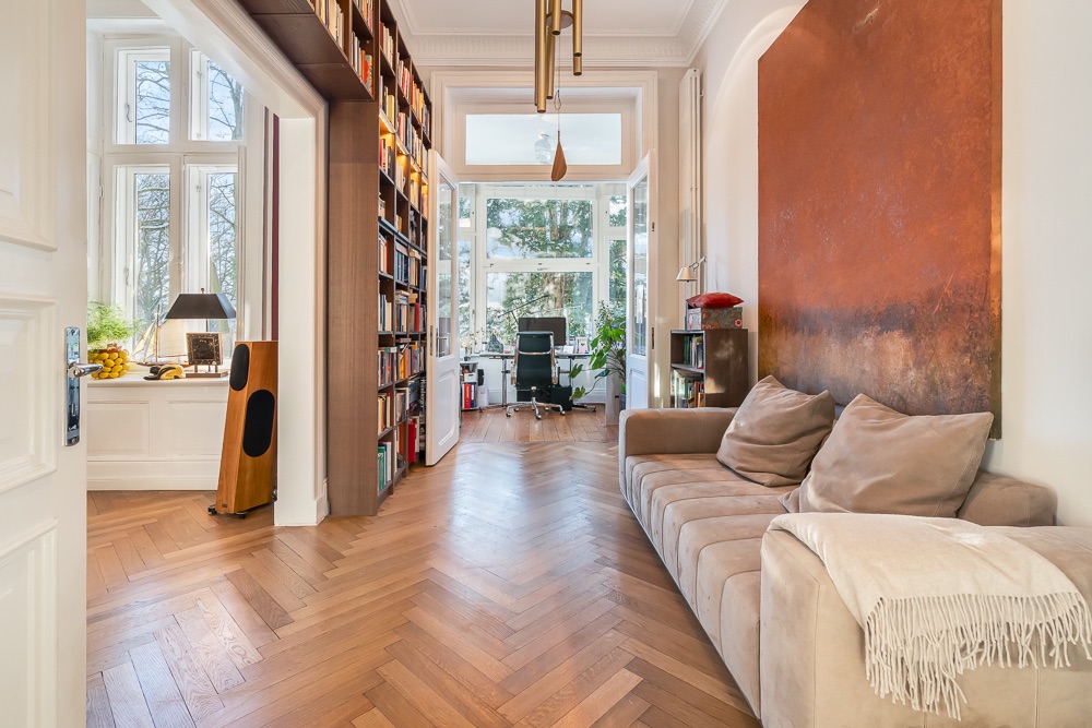 Helles Wohnzimmer mit Sofa, Bücherregal und Schreibtisch am Fenster, Parkettboden.