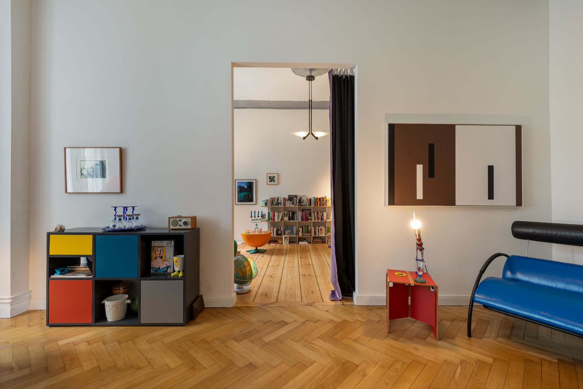 Wohnzimmer mit bunter Bank, Regal, Kunst an der Wand und Blick in ein weiteres Zimmer mit Bücherregal.