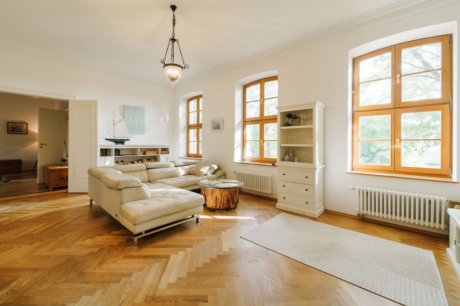 Helles Wohnzimmer mit Ecksofa, Holzboden, großen Fenstern und Regal.