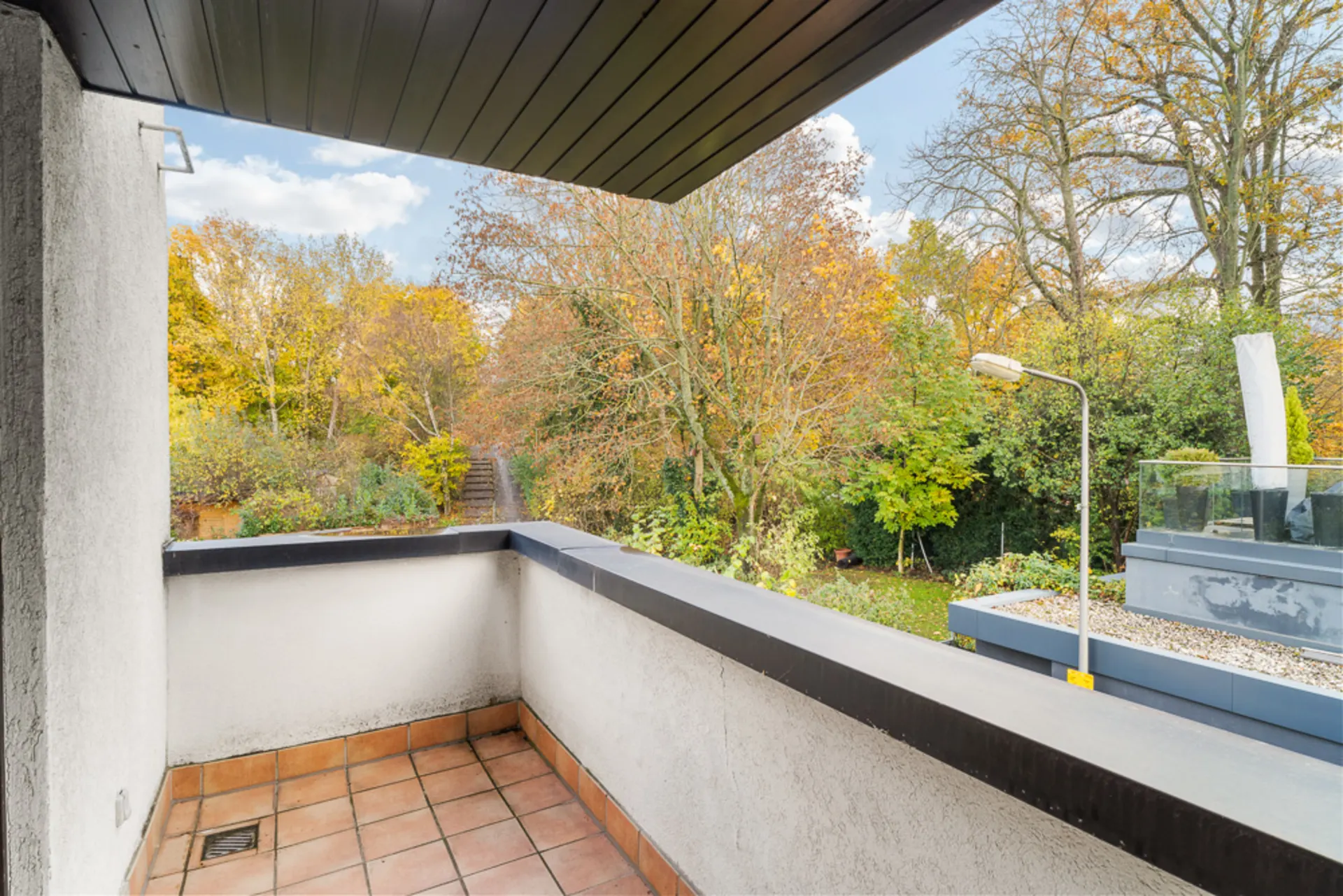 Balkon mit Fliesenboden und Blick auf einen grünen Garten mit Bäumen im Herbst.