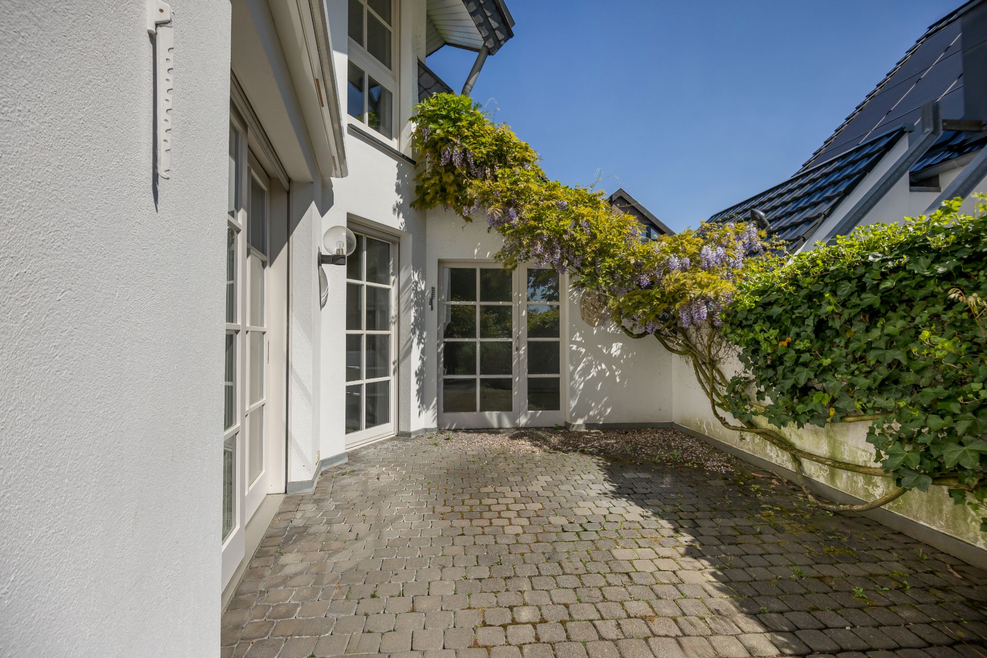Gepflasterte Terrasse mit weißer Hauswand, vielen Fenstern und blühender Kletterpflanze am Zaun.