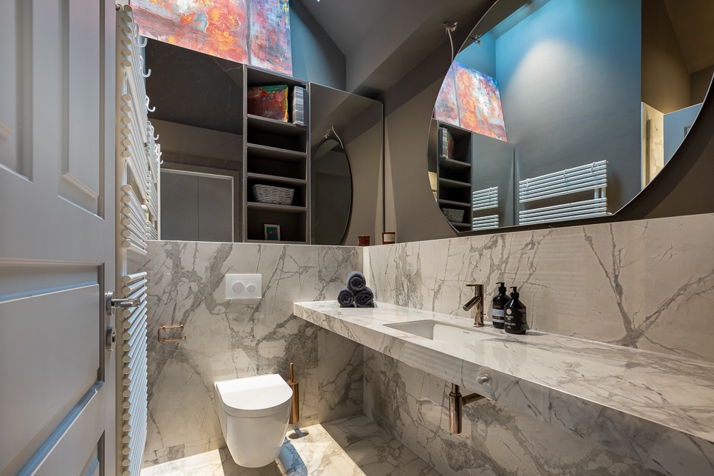 Modernes Badezimmer mit Marmorausstattung, rundem Spiegel, Waschbecken und Wandtoilette.