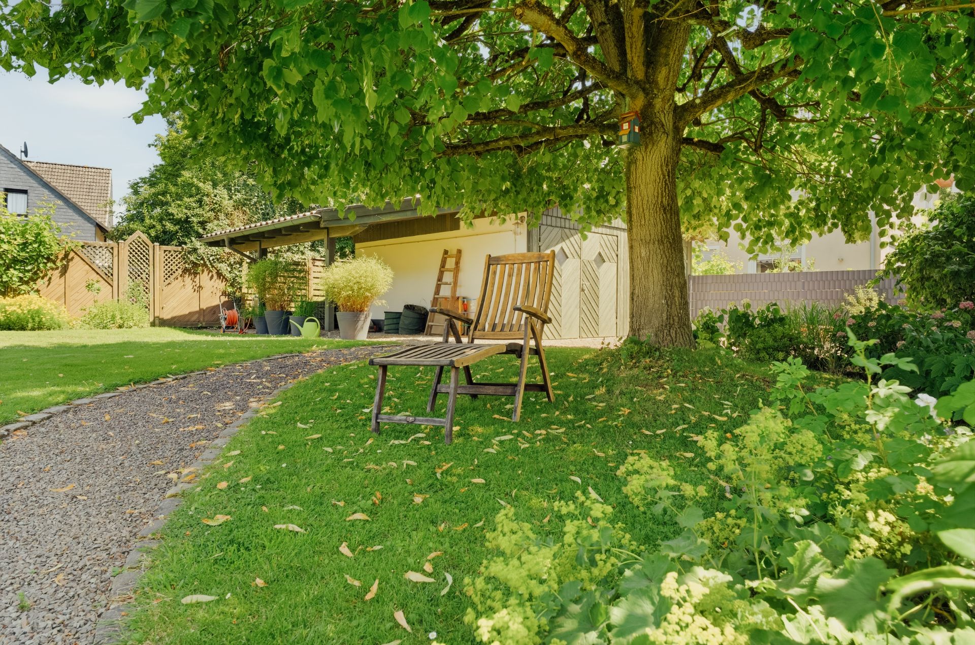 Garten mit Kiesweg, großer Baum, zwei Holzstühlen und Tisch im Schatten, im Hintergrund ein Schuppen.