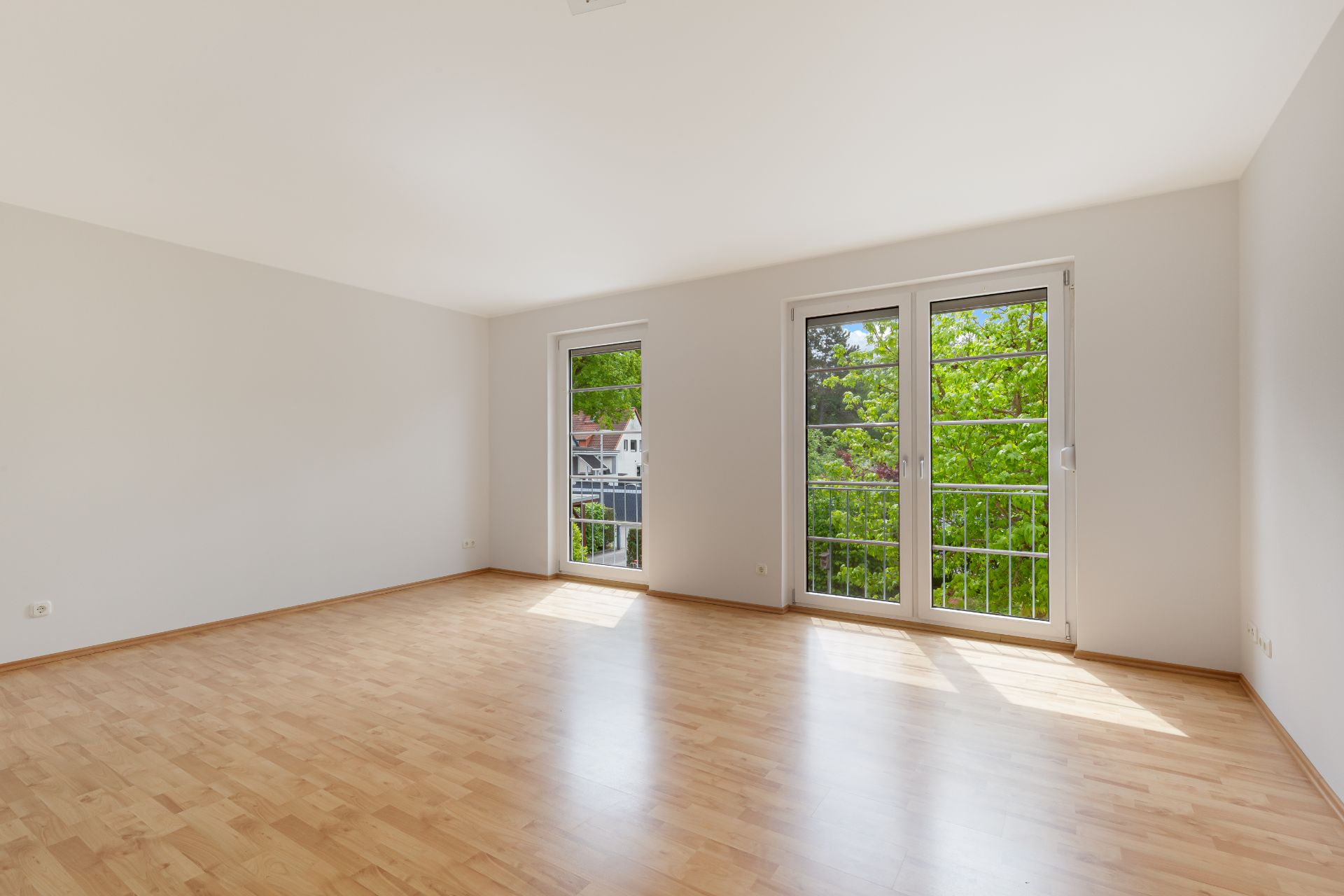 Heller, leerer Raum mit Parkettboden und großen Fenstern mit Blick ins Grüne.