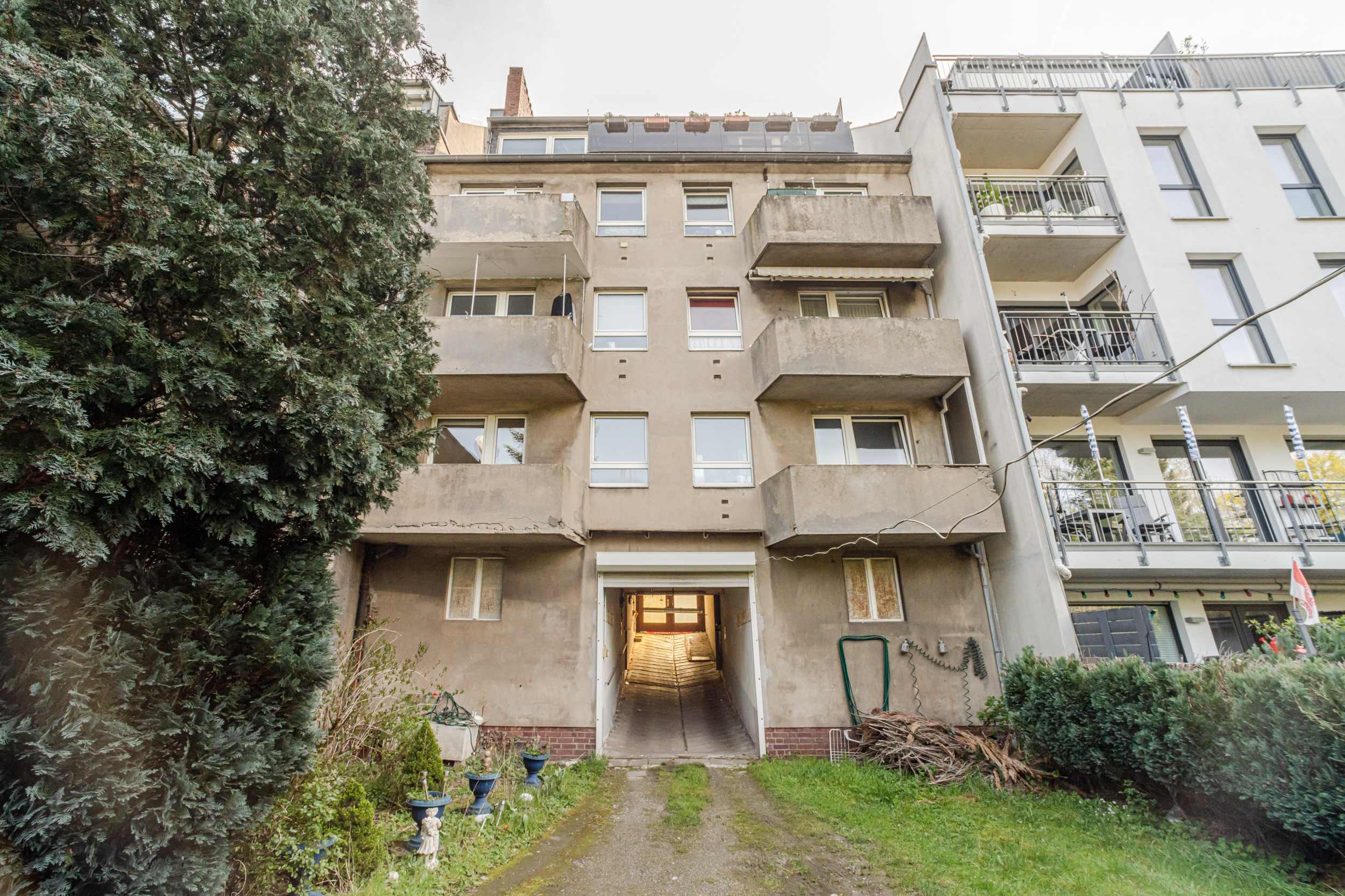 Mehrstöckiges Wohnhaus mit Balkonen und Durchfahrt, umgeben von Bäumen und Sträuchern.