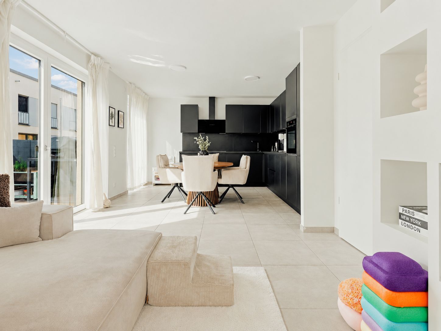 Helles Wohnzimmer mit Sofa, Essbereich und moderner schwarzer Küche, große Fenster mit Vorhängen.