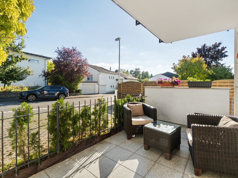 Terrasse mit zwei Korbstühlen, Tisch und Blumen, Blick auf Wohnstraße und Nachbarhäuser.