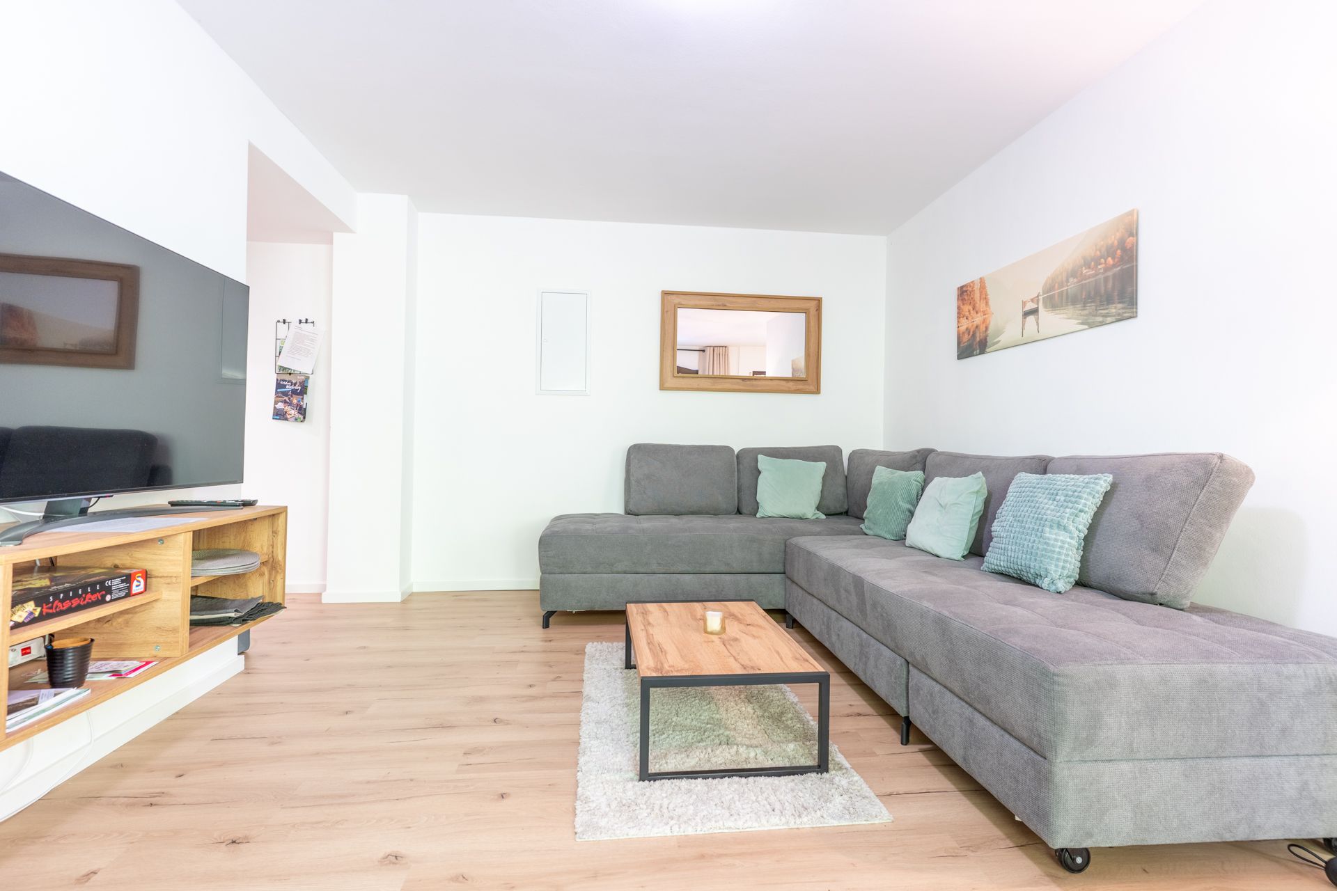 Helles Wohnzimmer mit grauem Ecksofa, Couchtisch und Fernseher auf Holzregal.