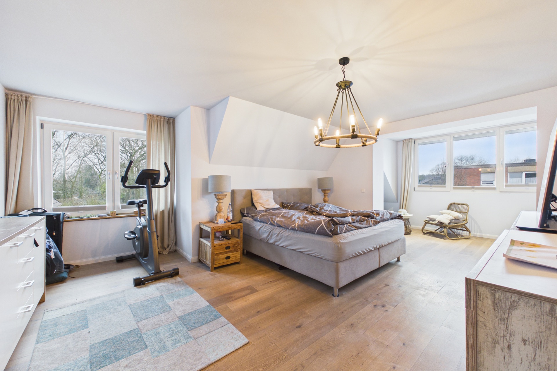Helles Schlafzimmer mit Doppelbett, Teppich, Heimtrainer, Kommode und großen Fenstern.