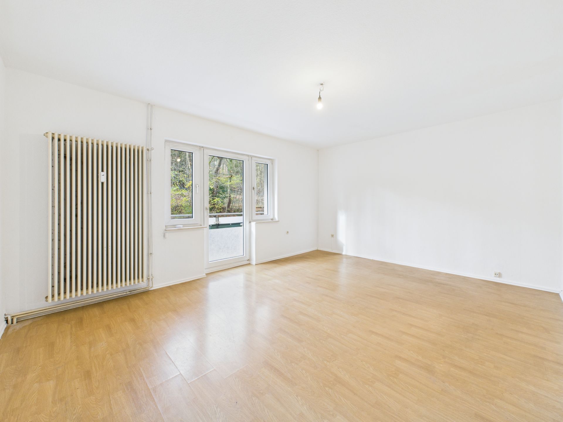 Helles, leeres Zimmer mit Parkettboden, großem Fenster und Heizung an der Wand.
