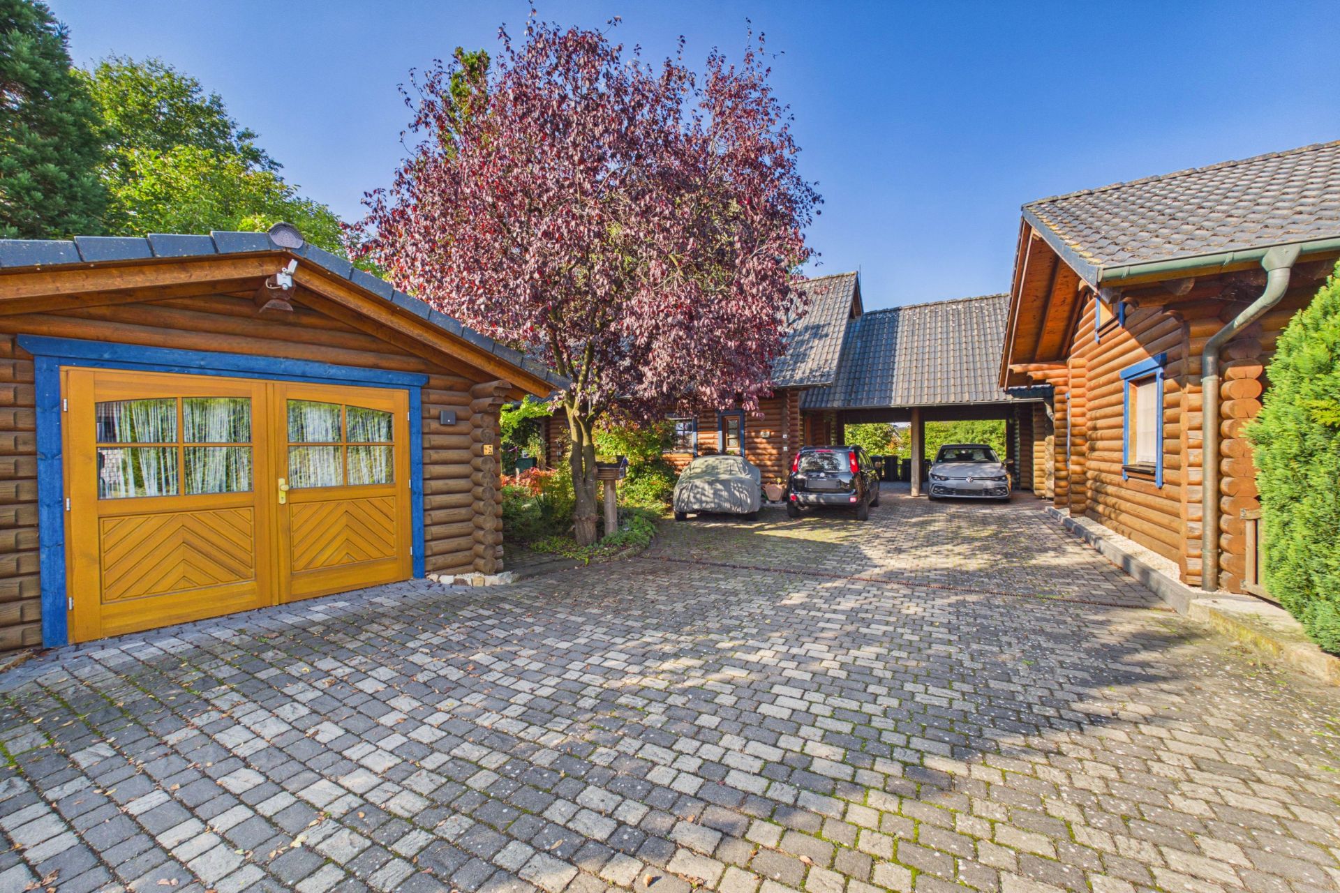 Blockhaus mit gelber Garage, gepflasterter Einfahrt und drei Autos vor dem Haus.