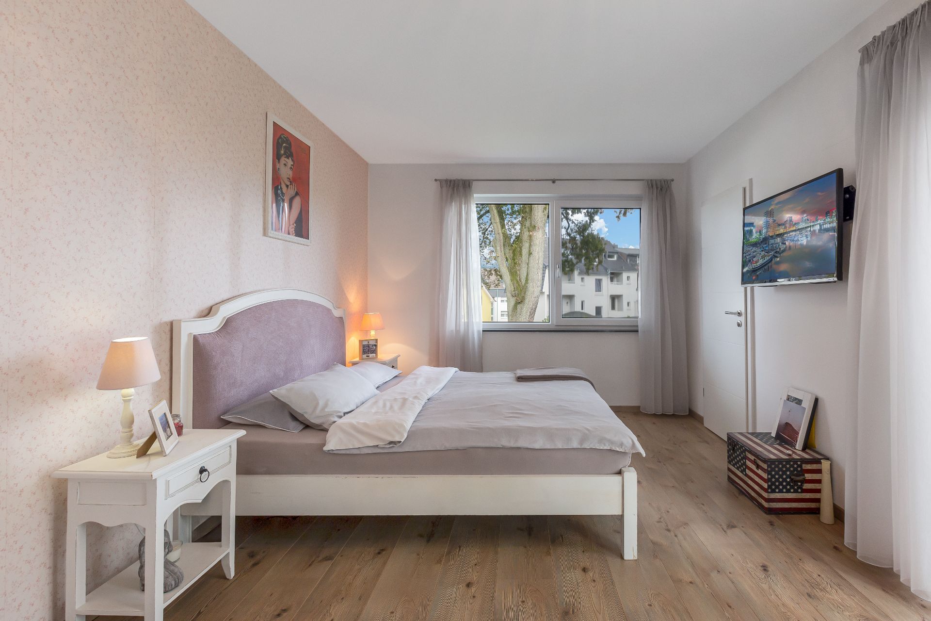 Helles Schlafzimmer mit Doppelbett, Nachttisch, Fenster und Wandfernseher.