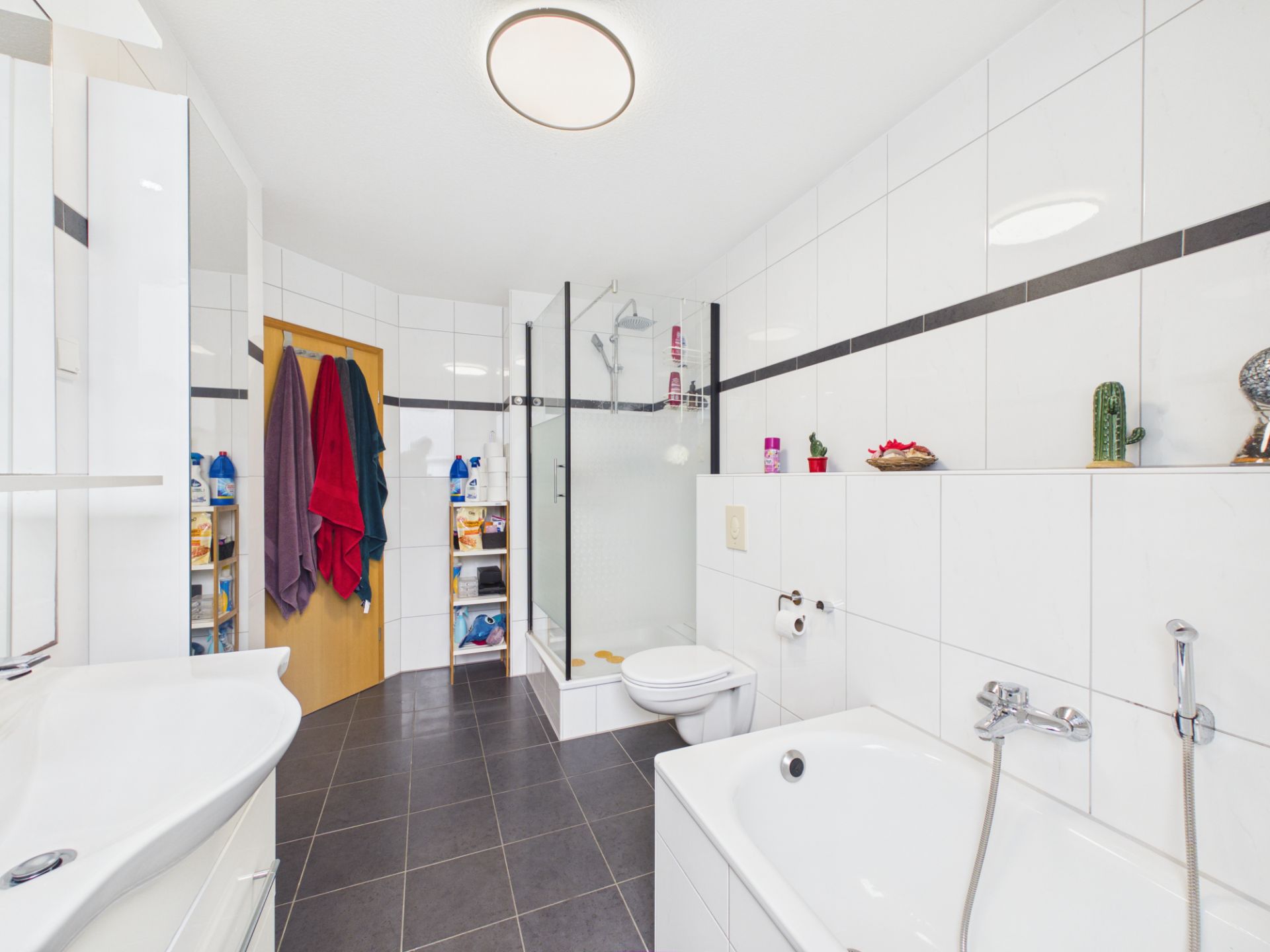 Modernes Badezimmer mit Badewanne, Dusche, Toilette und Regalen, helle Fliesen und dunkler Boden.