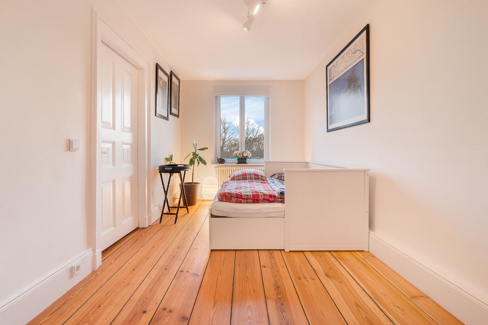 Helles Schlafzimmer mit Holzboden, Einzelbett, Pflanzen und großem Fenster.