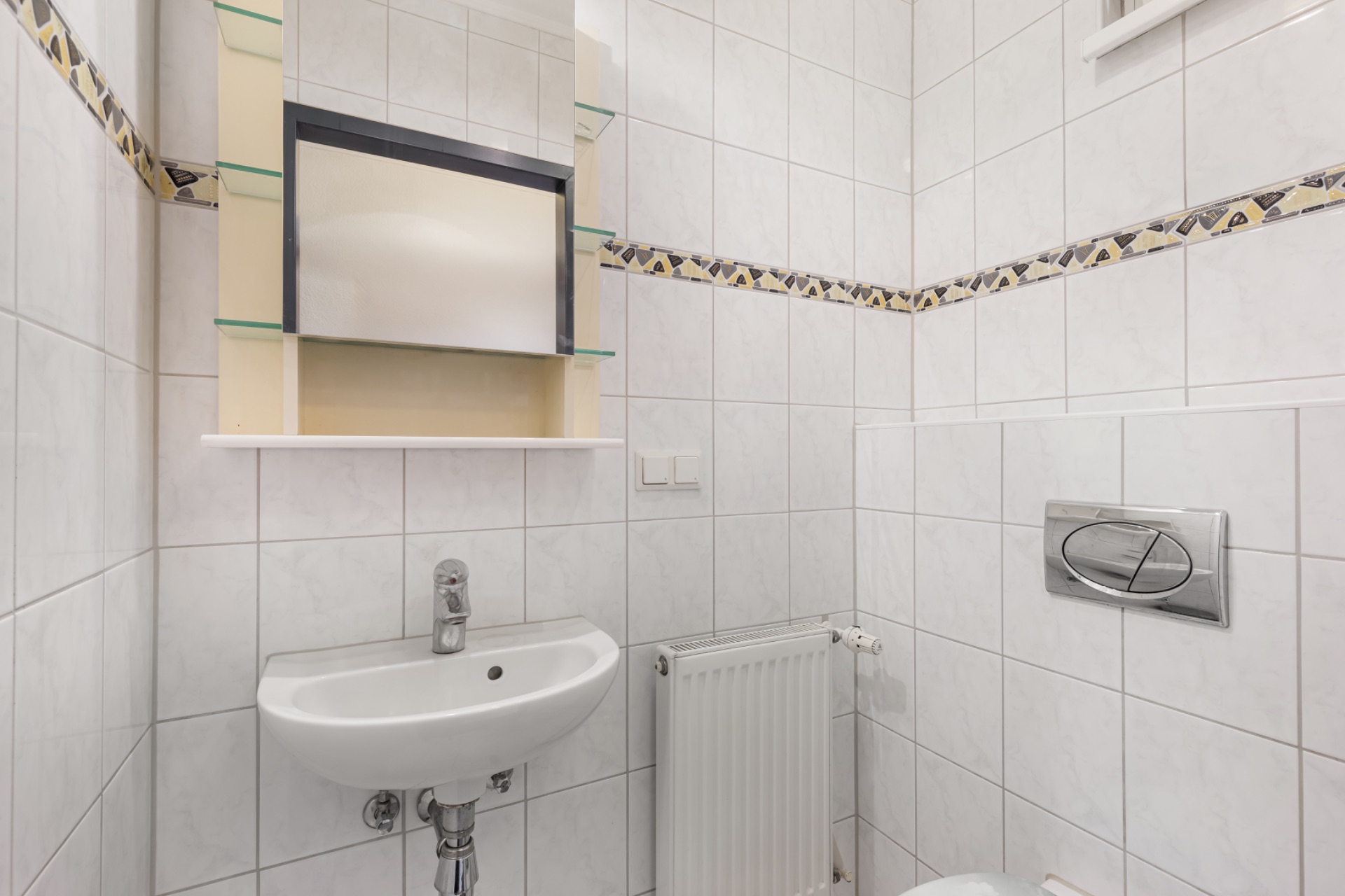 Kleines Badezimmer mit Waschbecken, Spiegel, Heizung und WC, weiße Fliesen an den Wänden.