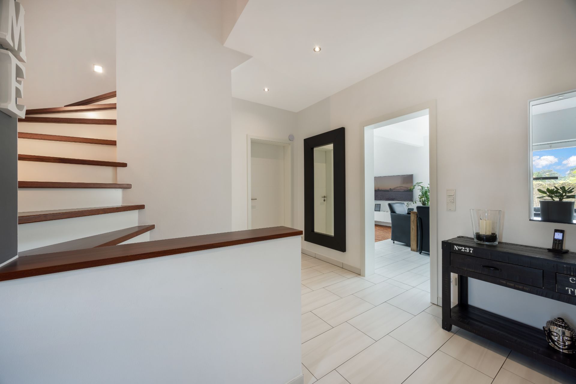 Heller Flur mit Treppe, Spiegel, Kommode und Blick in ein Wohnzimmer.