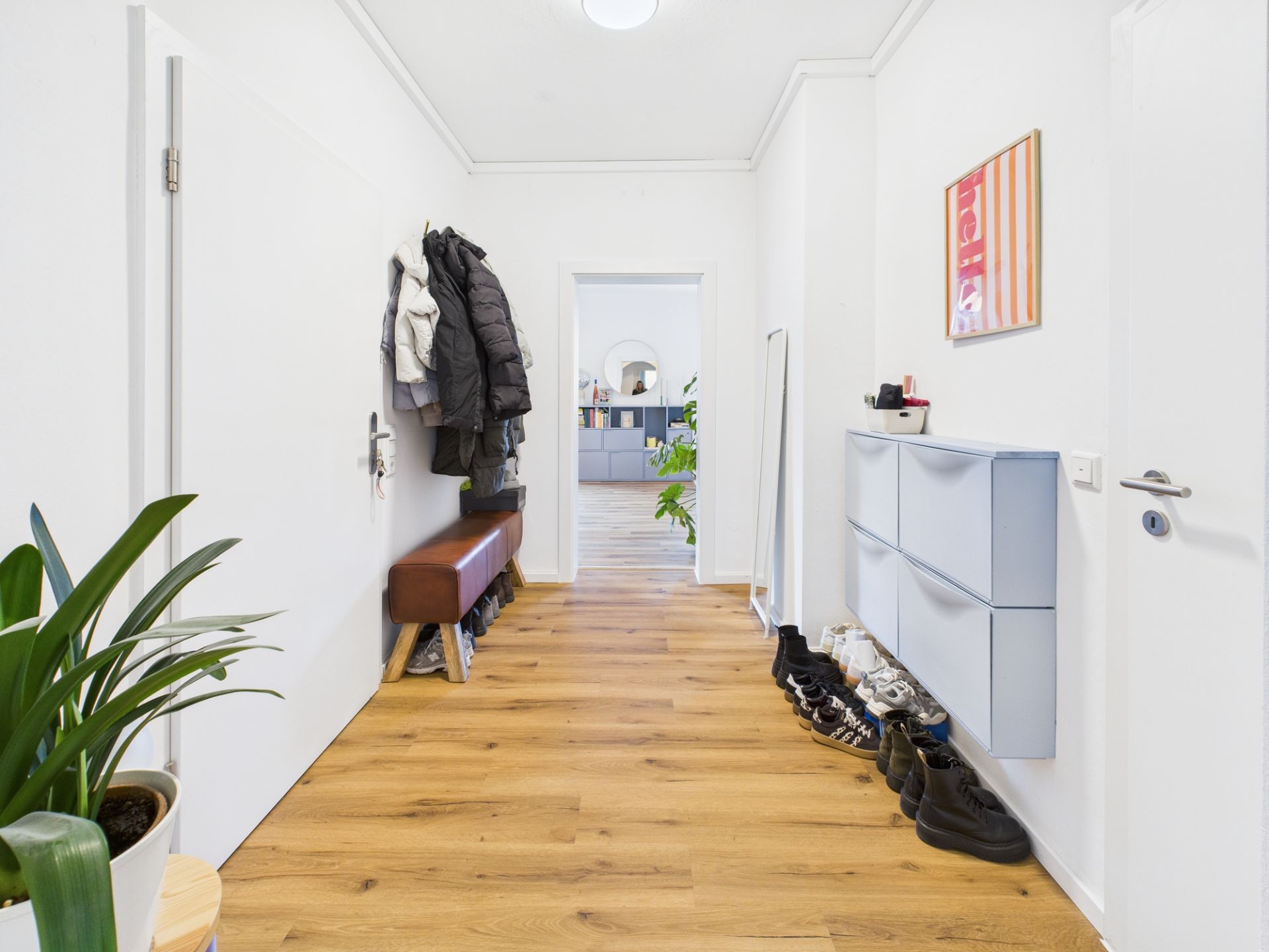 Flur mit Holzboden, Schuhregal, Jacken, Bank und Pflanze, Blick ins Wohnzimmer.