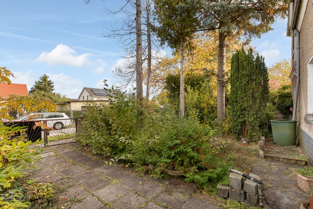 Gepflasterter Gartenbereich mit Bäumen, Sträuchern und einer grünen Regentonne neben einem Haus.