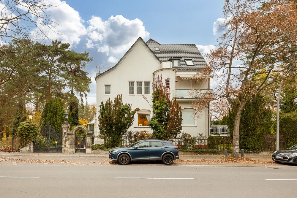 Weiße Wohnvilla mit Balkon, Auto davor und Bäumen an der Straße.