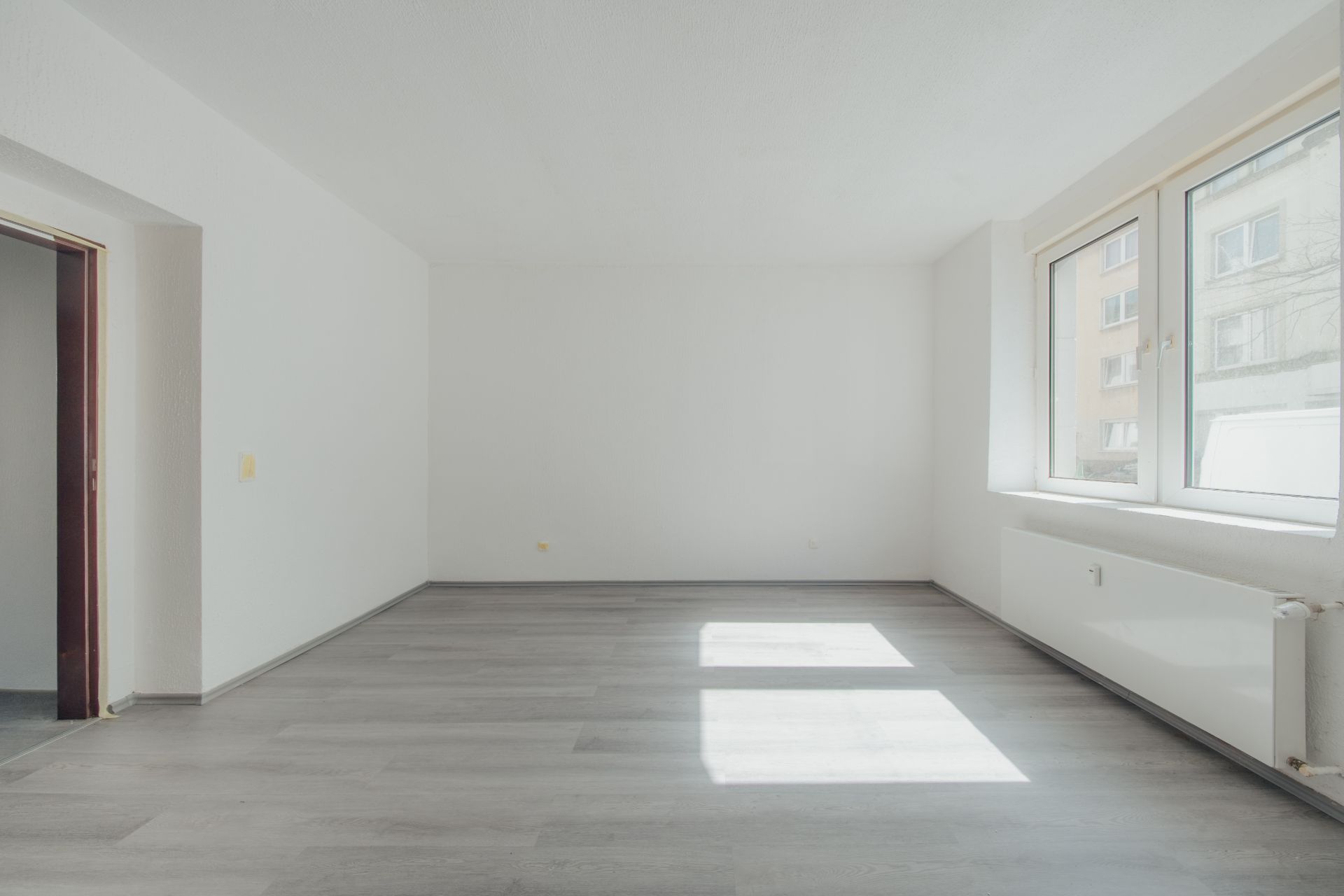 Heller, leerer Raum mit weißen Wänden, großem Fenster und grauem Boden.