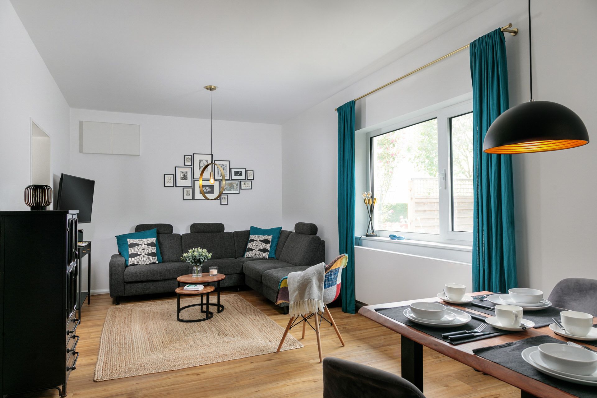 Helles Wohnzimmer mit Ecksofa, Esstisch, großem Fenster und türkisfarbenen Vorhängen.