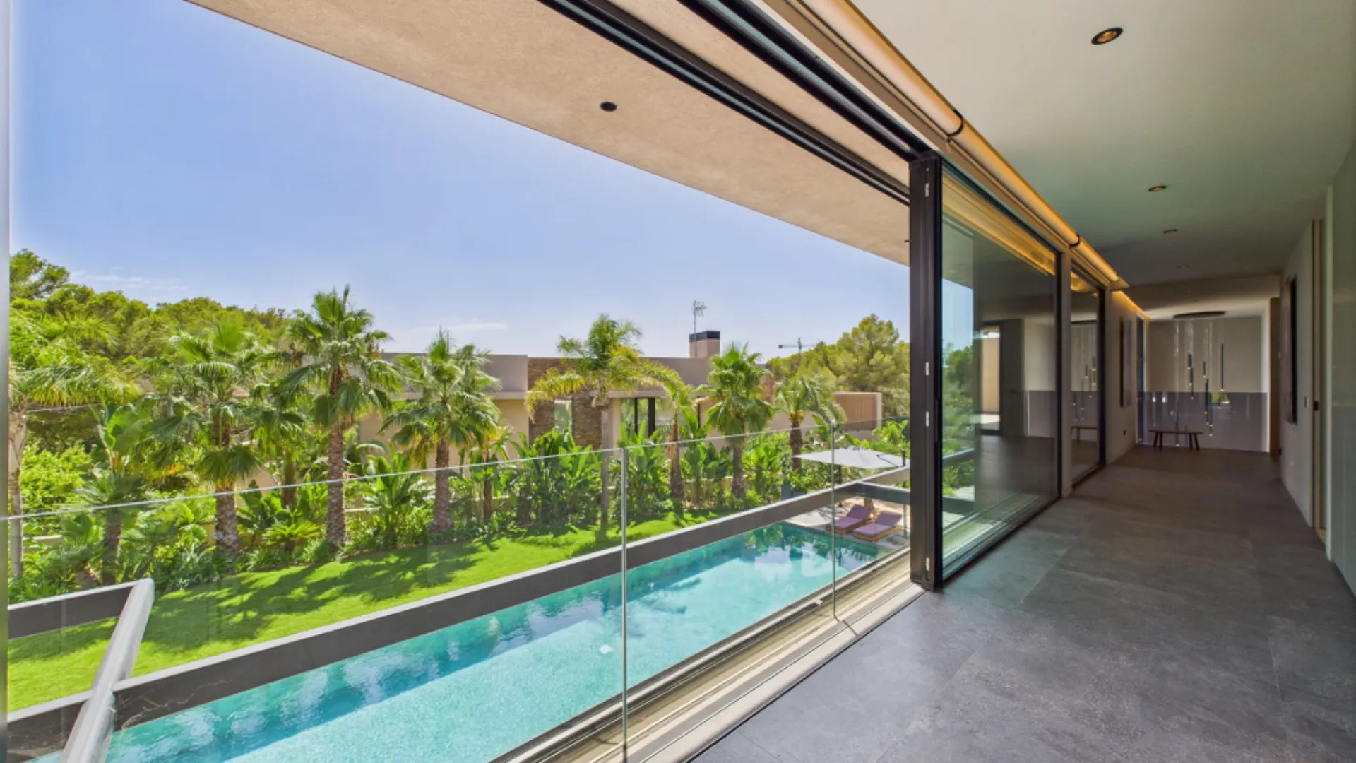 Großer Balkon mit Glasgeländer, Blick auf Pool und Palmen im Garten.
