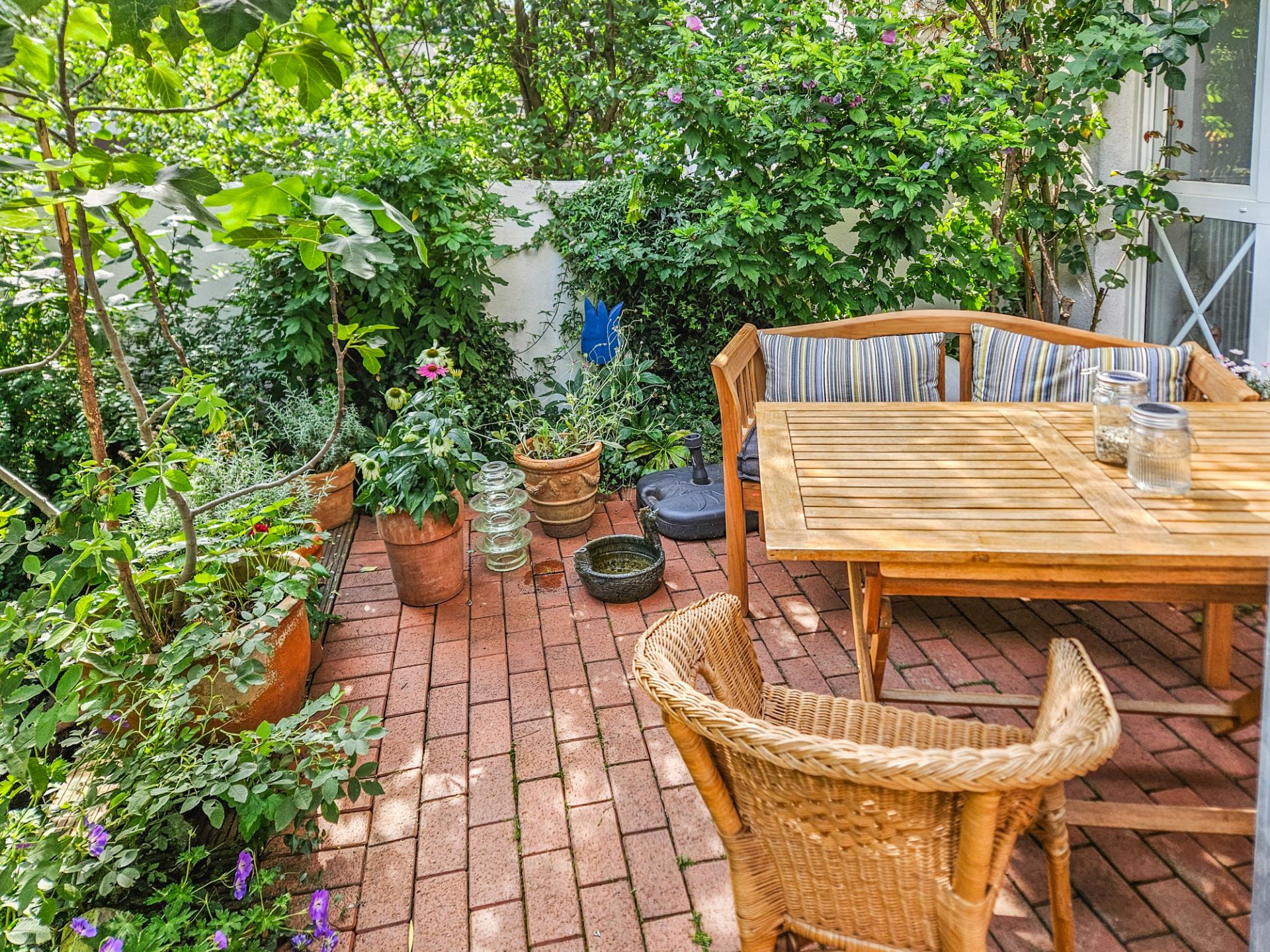 Kleiner Garten mit Holzsitzgruppe, Ziegelboden und vielen grünen Pflanzen in Töpfen.
