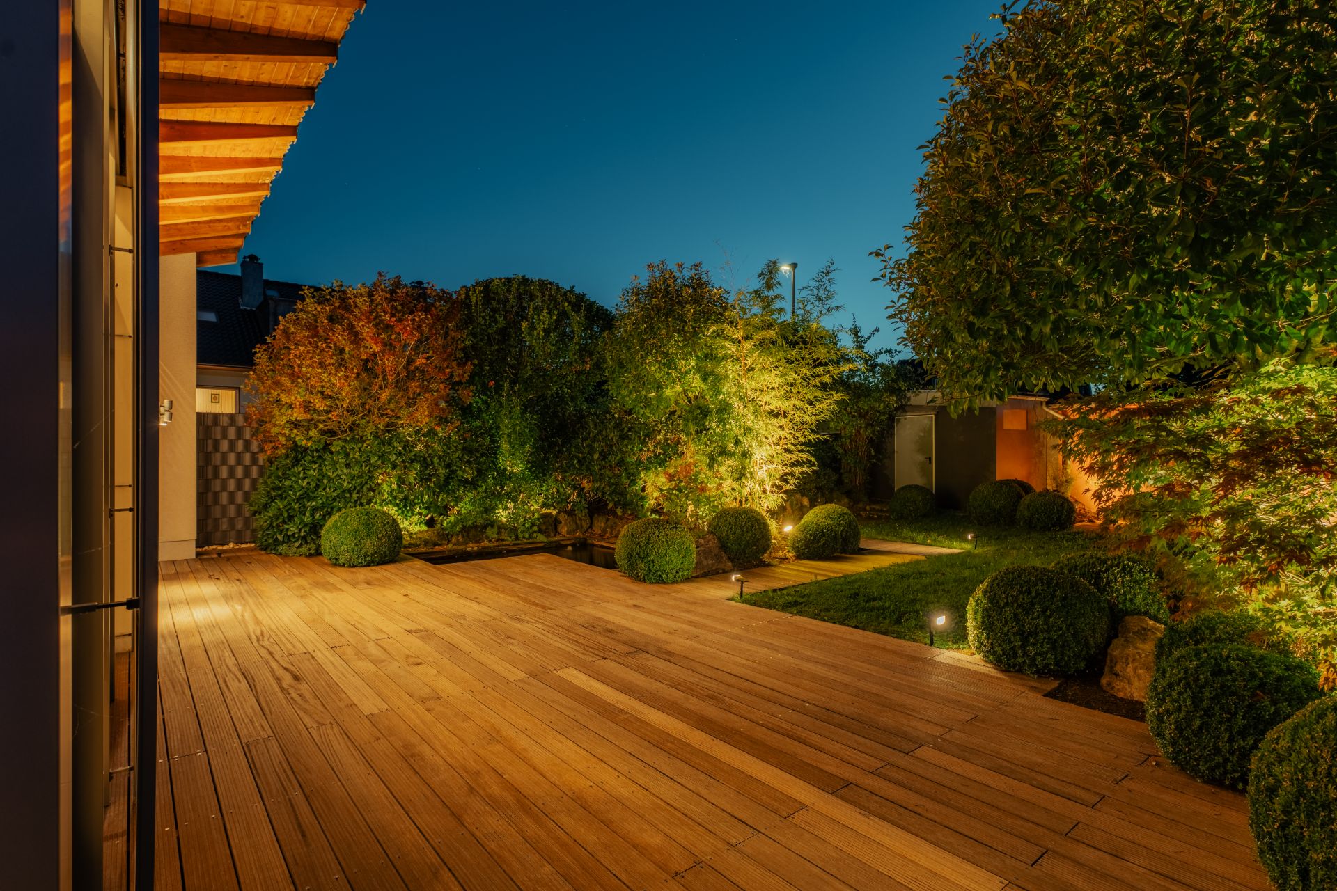 Holzterrasse mit Garten und beleuchteten Büschen am Abend.