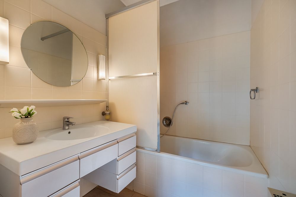 Helles Badezimmer mit Waschtisch, rundem Spiegel und Badewanne mit Duschtrennwand.