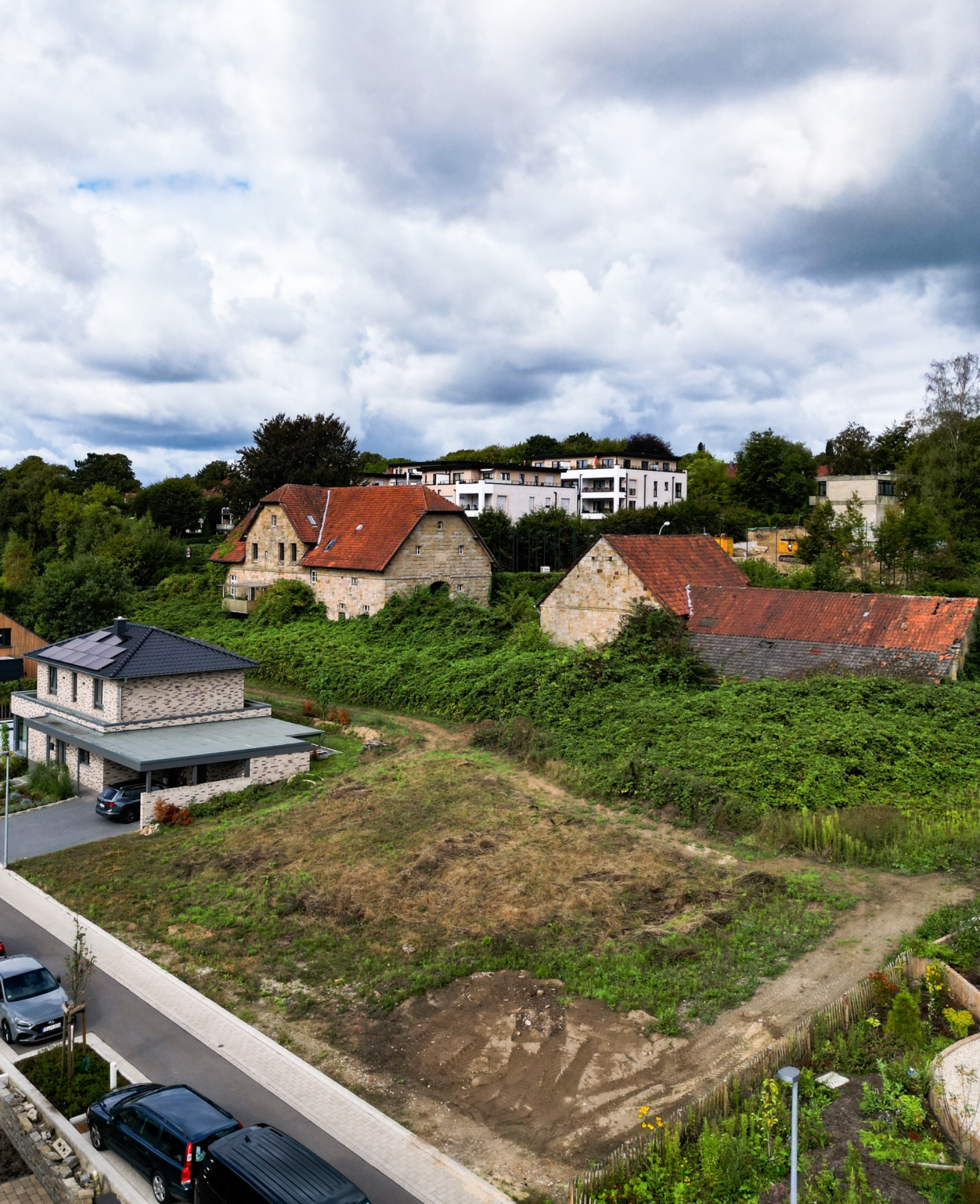 Blick auf ein leeres Grundstück neben Wohnhäusern mit alten Gebäuden und bewachsener Umgebung.