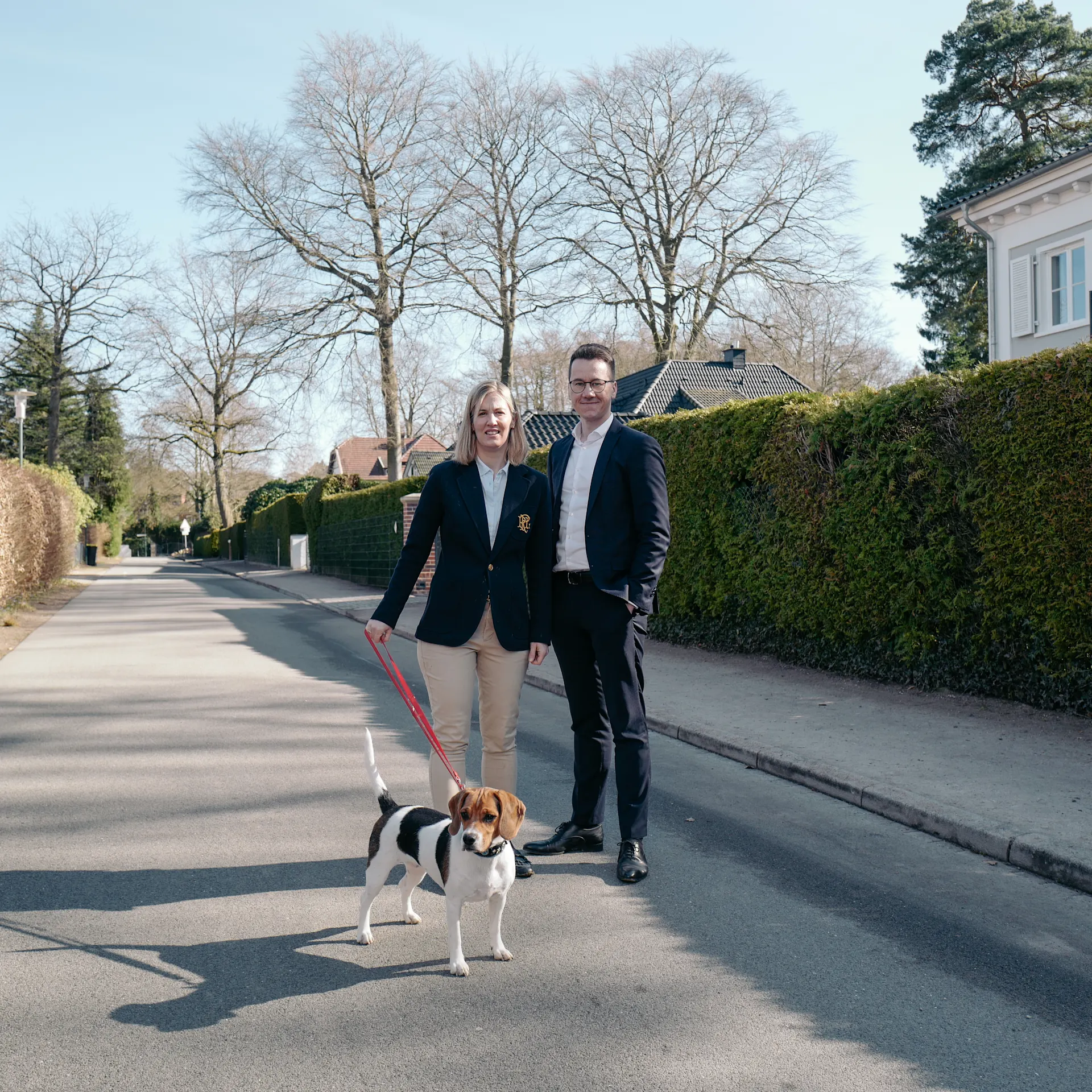 Zwei Personen mit einem Hund an der Leine stehen auf einer ruhigen Straße vor Wohnhäusern.