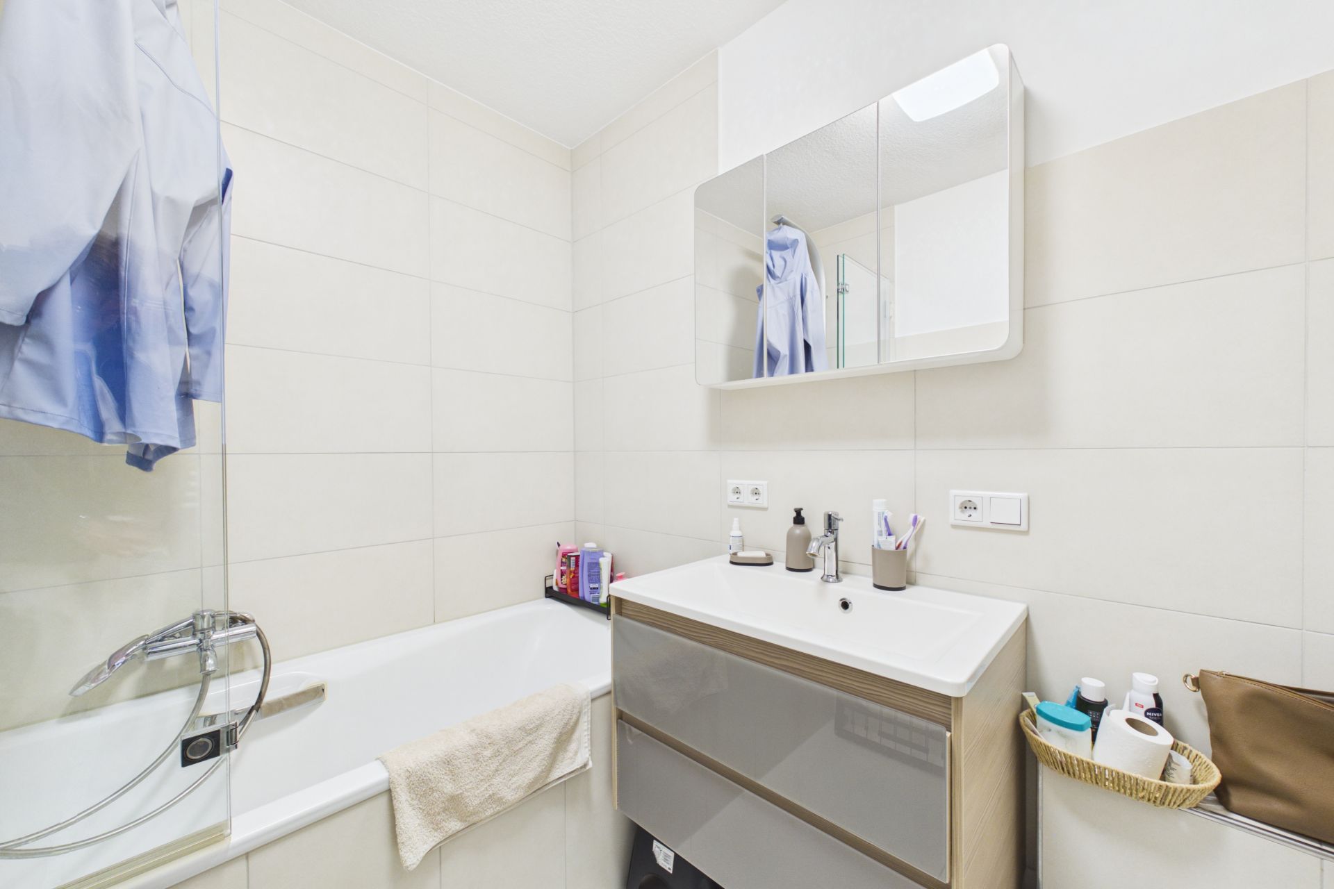 Helles Badezimmer mit Badewanne, Waschbecken, Spiegel und Ablagen mit Pflegeprodukten.